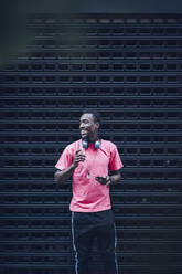 Porträt eines lächelnden Mannes mit Kopfhörern und Smartphone und rosa T-Shirt - OCMF00520