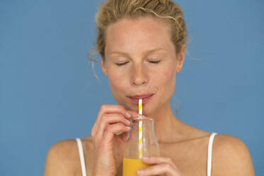 Blond woman with closed eyes drinking a juice, blue background - JOSF03553
