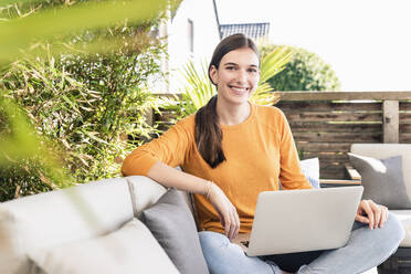 Porträt einer lächelnden jungen Frau, die mit einem Laptop auf einer Terrasse sitzt - UUF18237