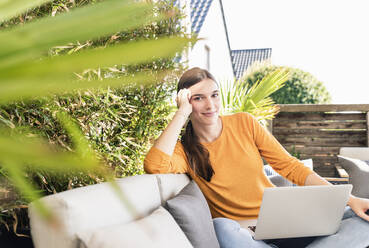 Porträt einer lächelnden jungen Frau, die mit einem Laptop auf einer Terrasse sitzt - UUF18238