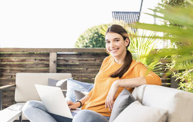 Porträt einer lächelnden jungen Frau, die mit einem Laptop auf einer Terrasse sitzt - UUF18239