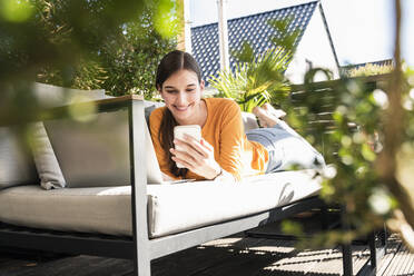 Junge Frau liegt auf einer Couch auf der Terrasse und benutzt Handy und Laptop - UUF18244