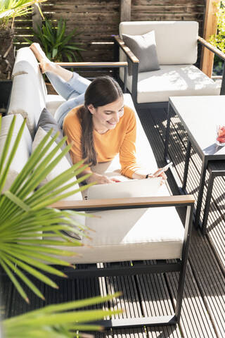 Junge Frau liegt auf einer Couch auf der Terrasse und benutzt Handy und Laptop, lizenzfreies Stockfoto