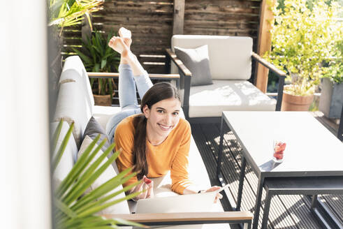 Porträt einer lächelnden jungen Frau, die auf einer Couch auf der Terrasse liegt und ein Mobiltelefon und einen Laptop benutzt - UUF18246