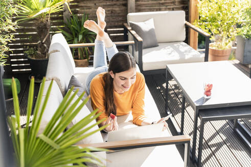 Junge Frau liegt auf einer Couch auf der Terrasse und benutzt Handy und Laptop - UUF18247