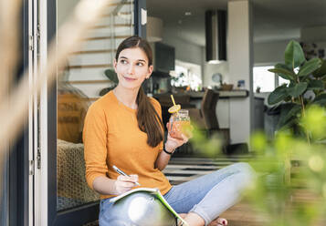 Young woman sitting with healthy drink and notebook at terrace door - UUF18253