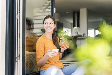 Smiling young woman sitting with healthy drink and cell phone at terrace door - UUF18254