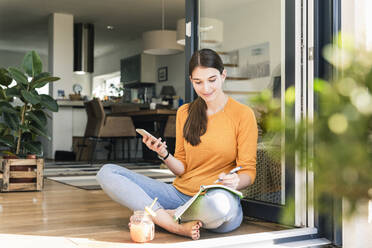 Young woman sitting with cell phone and notebook at terrace door - UUF18256