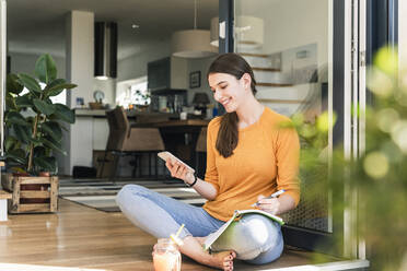 Young woman sitting with cell phone and notebook at terrace door - UUF18257