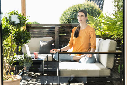 Young woman sitting on couch on terrace practicing yoga - UUF18260