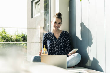 Smiling young woman with laptop, cell phone and healthy drink at home - UUF18272