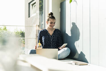 Smiling young woman with laptop, cell phone and healthy drink at home - UUF18273
