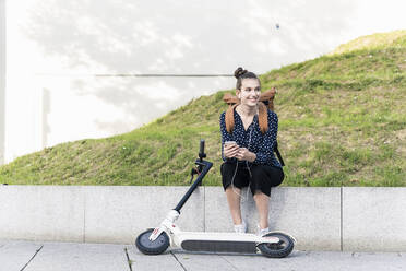 Smiling young woman with electric scooter, earphones and cell phone having a break - UUF18285