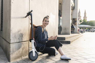 Lächelnde junge Frau mit Elektroroller, Kopfhörern und Mobiltelefon bei einer Pause in der Stadt - UUF18296