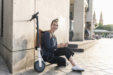Smiling young woman with electric scooter, earphones and cell phone having a break in the city - UUF18296