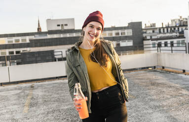 Porträt einer glücklichen jungen Frau mit Kopfhörern und Getränk auf einem Parkdeck - UUF18330