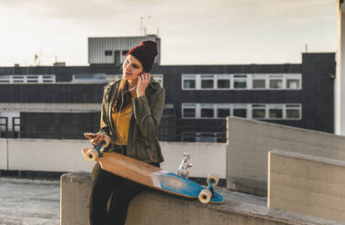 Stylische junge Frau mit Skateboard, Kopfhörern und Handy auf einem Parkdeck - UUF18335