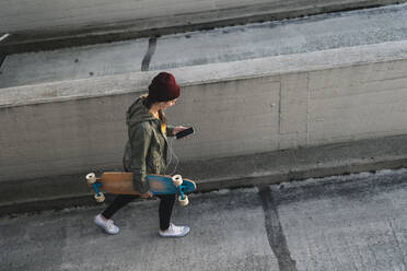 Stylische junge Frau mit Skateboard und Handy auf dem Parkdeck - UUF18343