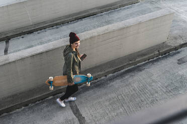 Stylische junge Frau mit Skateboard und Handy auf dem Parkdeck - UUF18344