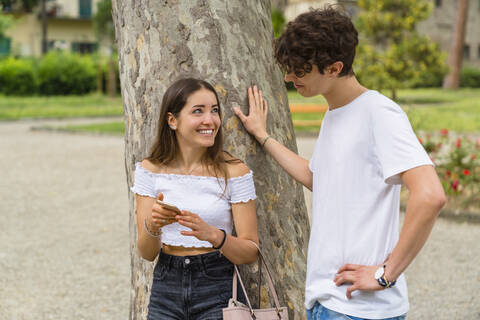 Junges Paar in einem Park, die Frau zeigt ihm ihr Smartphone und lächelt, lizenzfreies Stockfoto