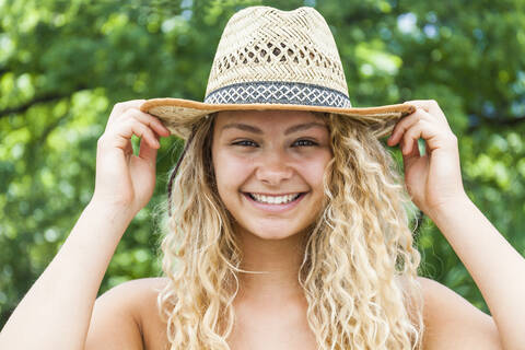 Porträt einer lächelnden blonden Frau mit Strohhut, Hände auf dem Hut, lizenzfreies Stockfoto