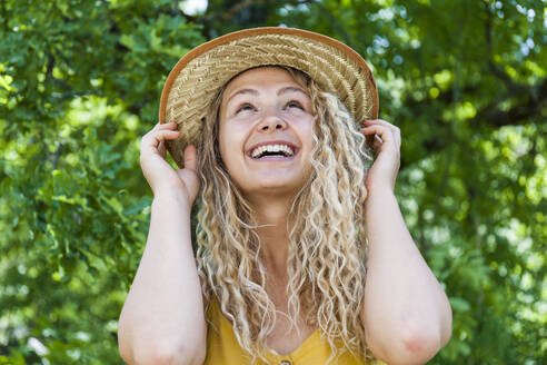 Porträt einer lächelnden blonden Frau mit Strohhut, Hände auf dem Hut - TCF06165