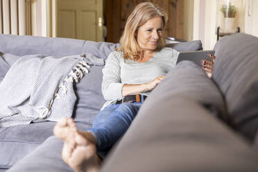 Lächelnde blonde Frau, die sich zu Hause auf der Couch entspannt und ein digitales Tablet benutzt - FMKF05756
