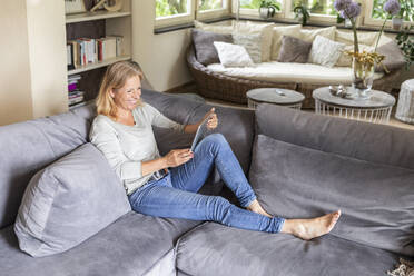 Laughing blond woman relaxing on couch at home using digital tablet - FMKF05757