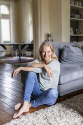 Portrait of smiling mature woman siiting barefoot on the floor in the living room - FMKF05759