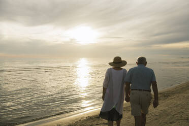 Rückenansicht eines älteren Paares, das Hand in Hand am Strand bei Sonnenuntergang spazieren geht, Liepaja, Lettland - AHSF00696