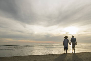 Back view of senior couple standing hand in hand on the beach watching sunset, Liepaja, Latvia - AHSF00698