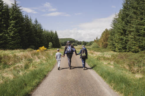 Rückenansicht eines Vaters, der Hand in Hand mit seinen Söhnen auf einer Landstraße spazieren geht, Cairngorms, Schottland, UK - NMS00328