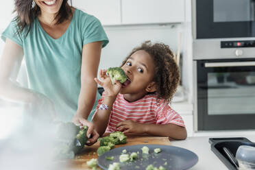 Girl cooking with mother in kitchen tasting broccoli - ERRF01667