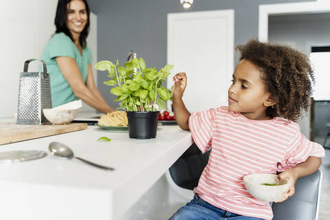 Mädchen kocht mit Mutter in der Küche und zupft Basilikumblätter, lizenzfreies Stockfoto