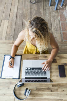 Junge Frau an einem hölzernen Schreibtisch, die einen Laptop benutzt und auf einem Klemmbrett schreibt - TCF06171