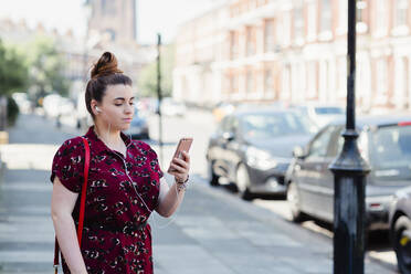 Portrait of woman with earphones standing on pavement looking at cell phone, Liverpool, UK - NMS00355