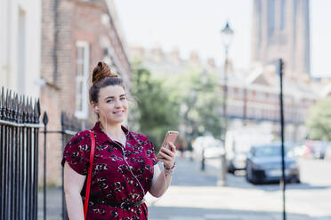 Portrait of woman standing on pavement using earphones and cell phone, Liverpool, UK - NMS00356