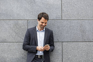 Smiling businessman with earphones looking at mobile phone - JUNF01711