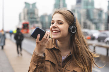 Glückliche junge Frau mit Kopfhörern und Mobiltelefon in der Stadt, London, UK - WPEF01652