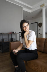 Smiling young woman on couch at home with cell phone - GIOF06918