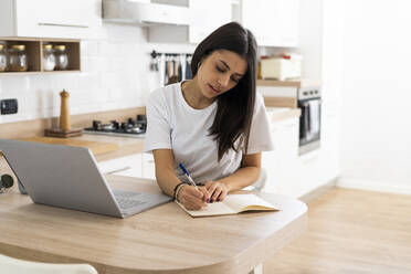 Junge Frau mit Laptop, die zu Hause Notizen macht - GIOF06928