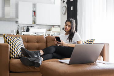 Young woman sitting on couch at home using laptop and cell phone - GIOF06936