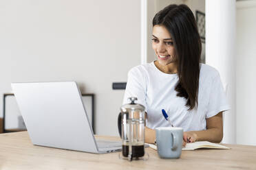 Junge Frau mit Laptop, die zu Hause Notizen macht - GIOF06939