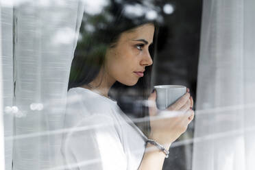 Junge Frau mit einer Tasse Kaffee hinter einer Fensterscheibe - GIOF06941