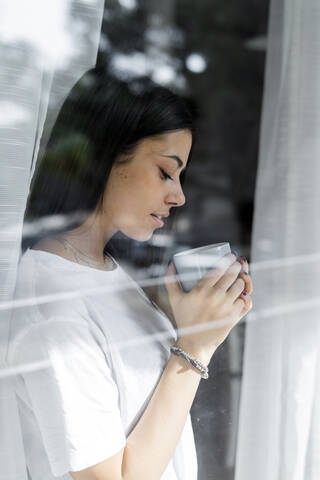 Junge Frau mit einer Tasse Kaffee hinter einer Fensterscheibe, lizenzfreies Stockfoto