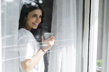 Lächelnde junge Frau mit einer Tasse Kaffee hinter einer Fensterscheibe - GIOF06943