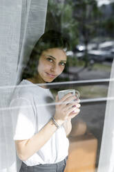 Porträt einer jungen Frau mit einer Tasse Kaffee hinter einer Fensterscheibe - GIOF06944