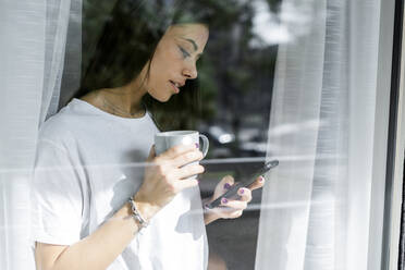 Junge Frau mit Kaffeetasse und Mobiltelefon hinter Fensterscheibe - GIOF06945