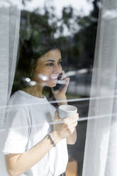 Lächelnde junge Frau am Mobiltelefon hinter einer Fensterscheibe - GIOF06946