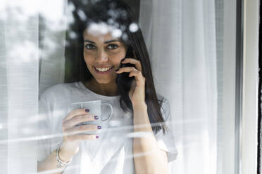 Porträt einer lächelnden jungen Frau am Handy hinter einer Fensterscheibe - GIOF06947